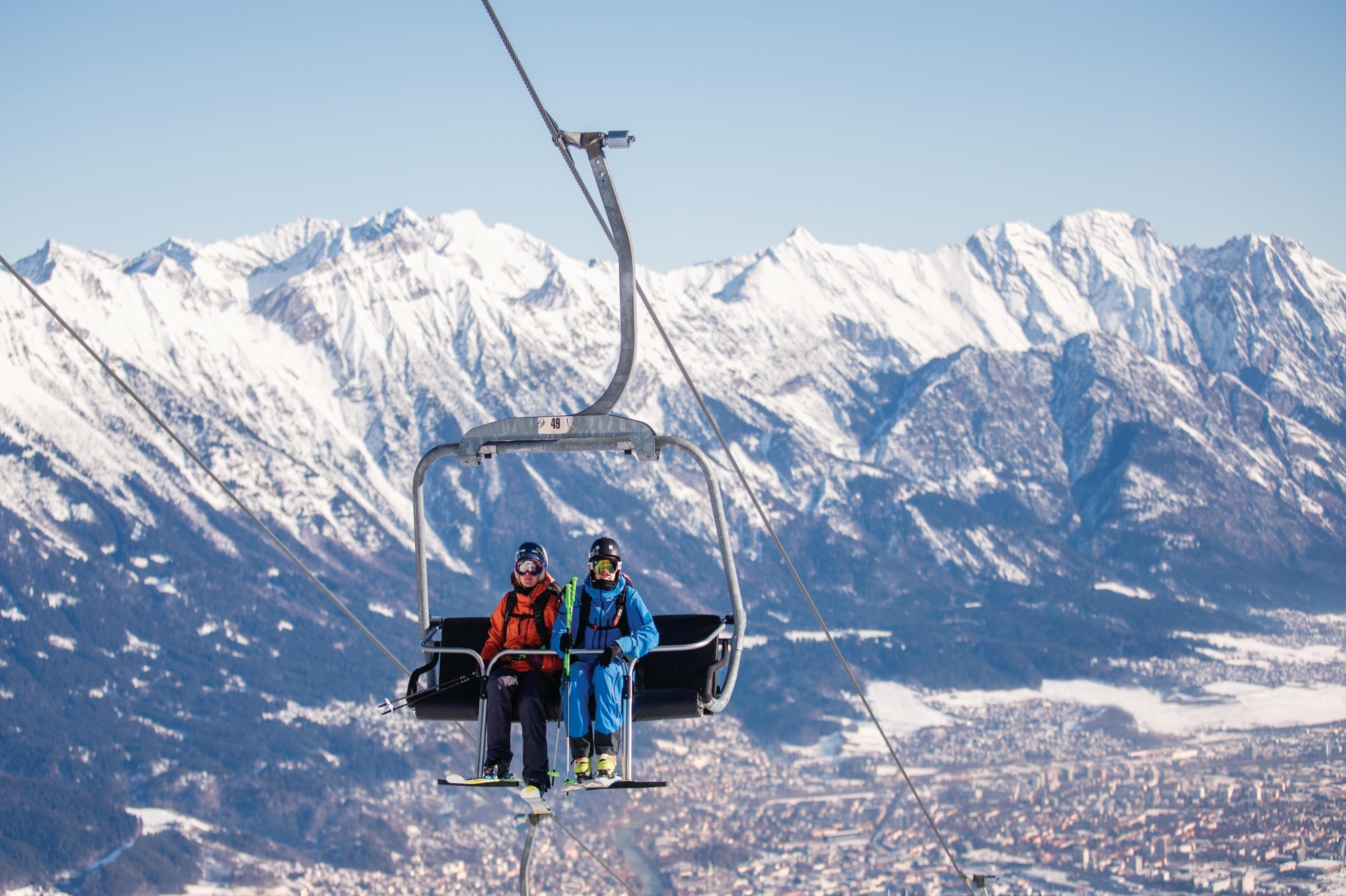 Zwei Skifahrer auf einem Sessellift mit beeindruckenden schneebedeckten Bergen im Hintergrund.